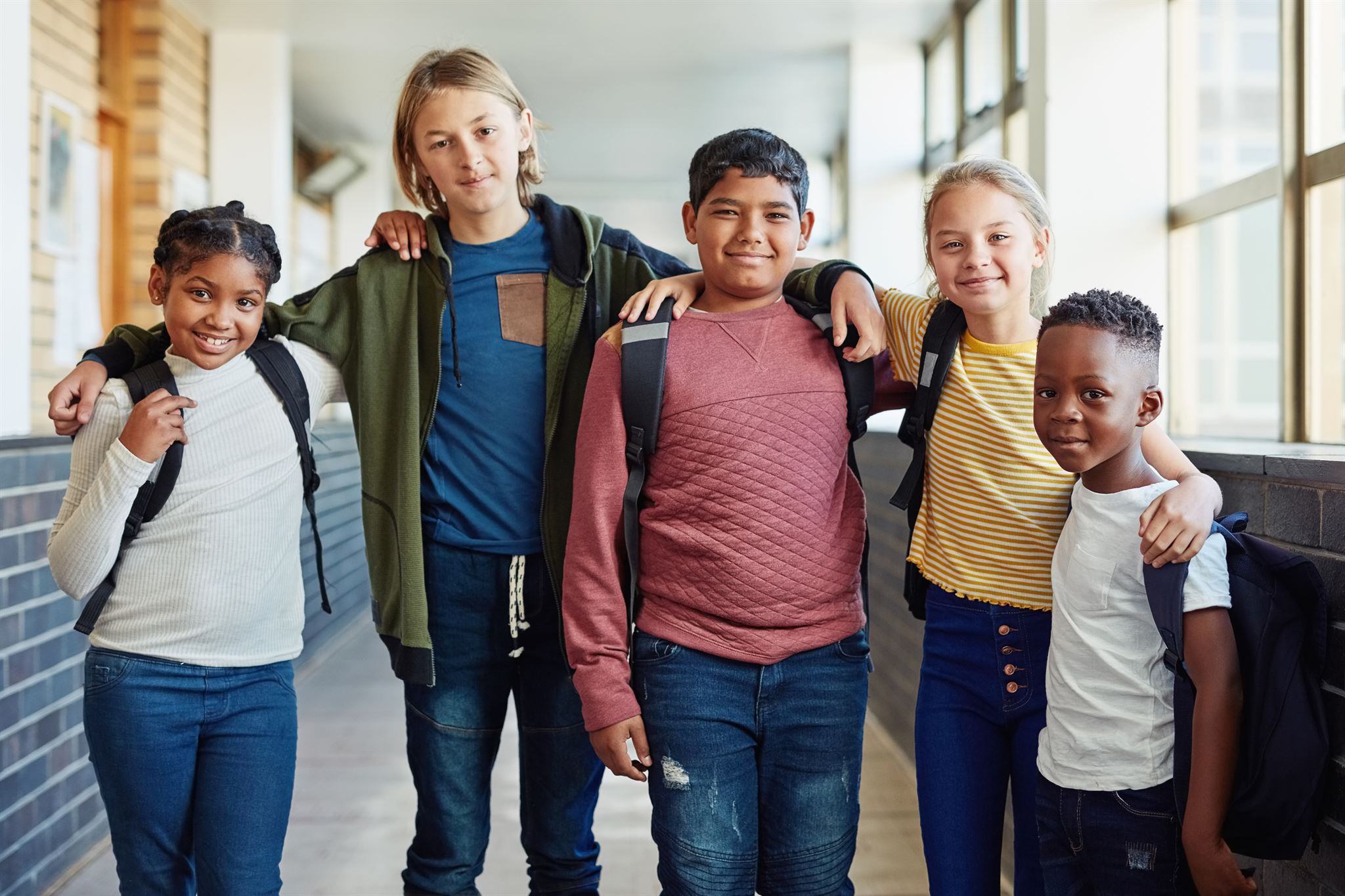 Portrait-of-five-young-children-with-arms-on-each-others-shoulders-standing-in-school-hallway